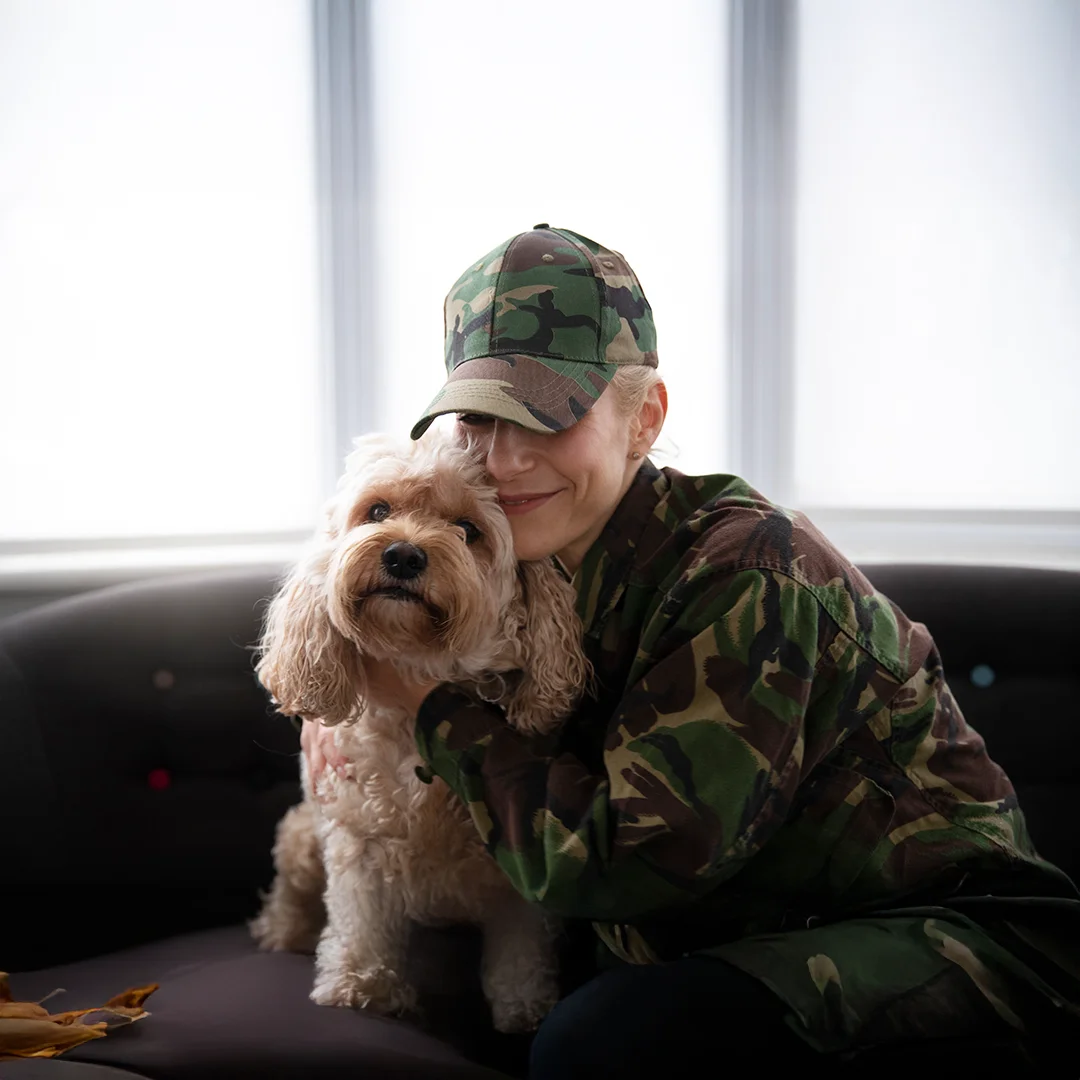 Military Woman Hugging Her Dog A woman in camouflage clothing lovingly hugs her fluffy tan dog while sitting on a couch near a bright window.